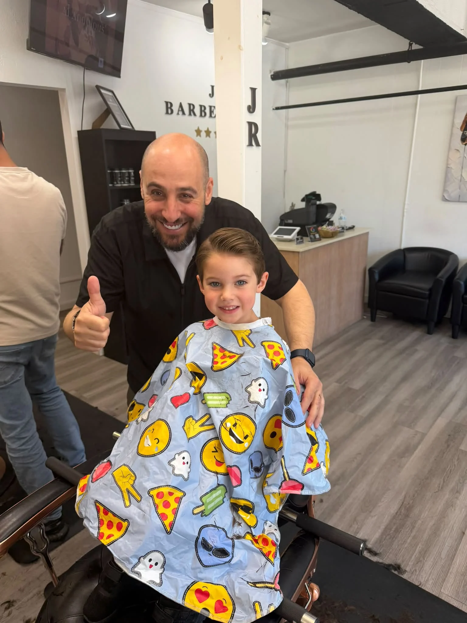 Barber and young customer smiling together beside a chair, with a colorful patterned cape