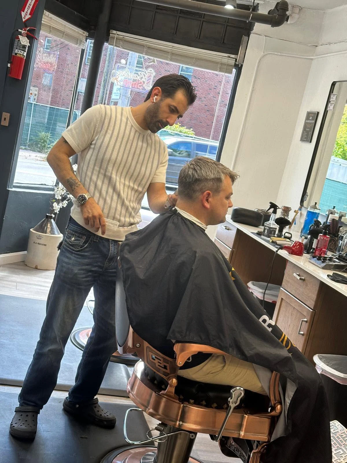 Barber working on a seated customer’s hair beside the front window