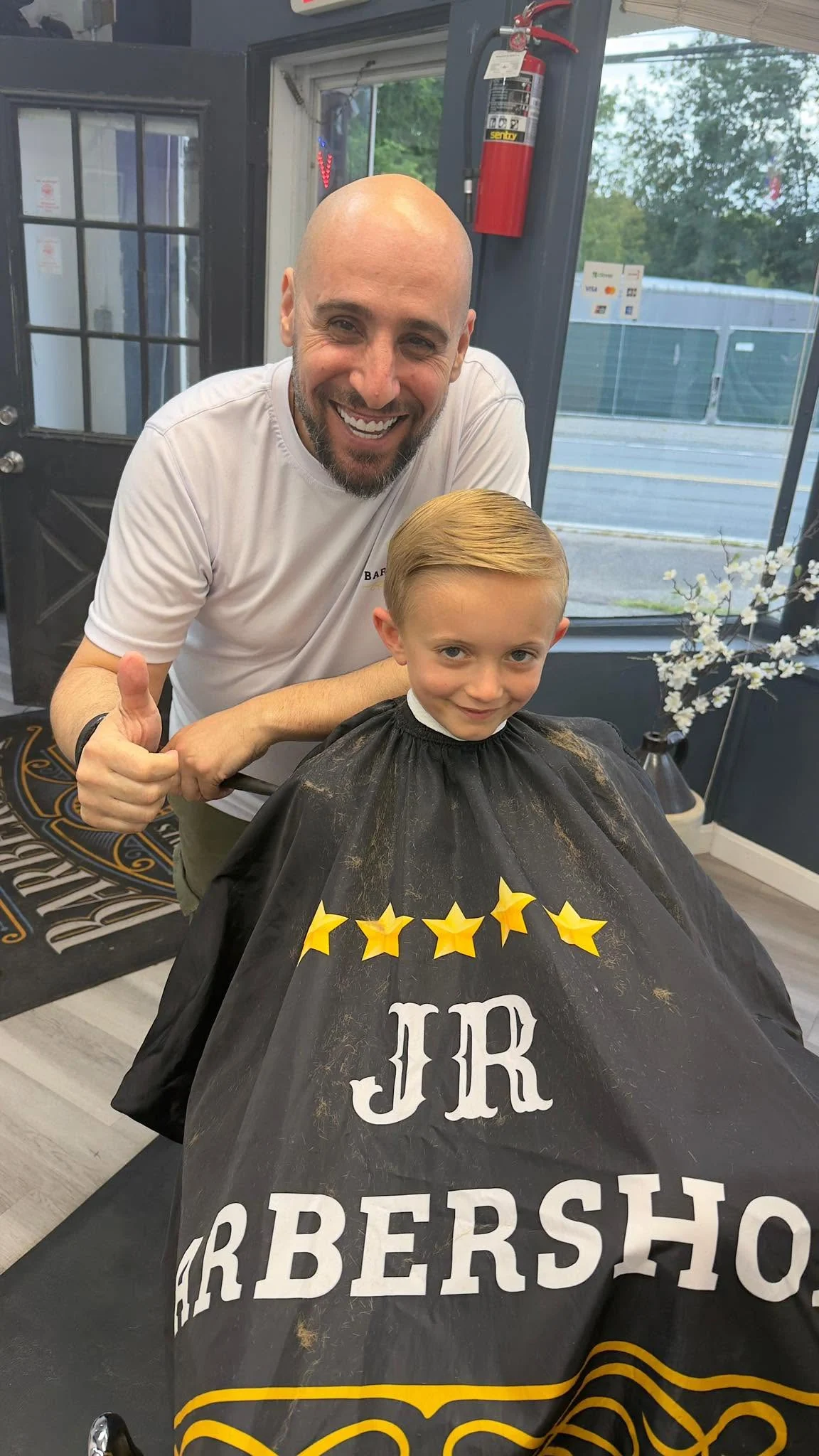 Barber giving a thumbs-up beside a young customer wearing a JR cape