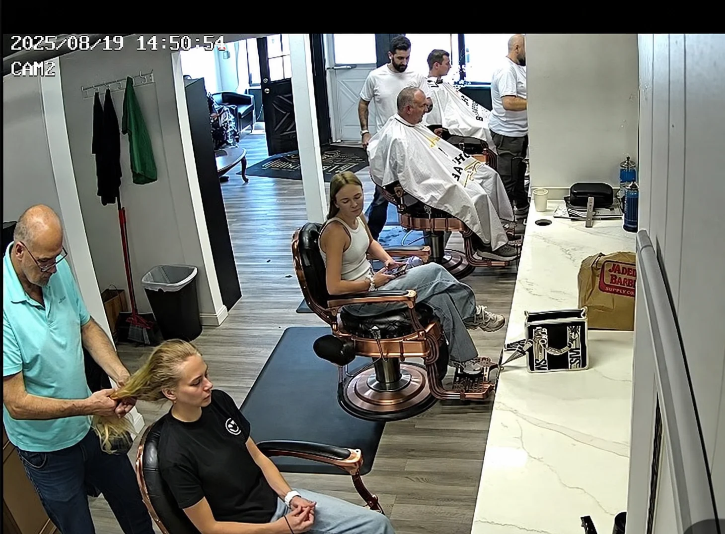 Overhead shop snapshot showing several customers and barbers, dated August 19, 2025