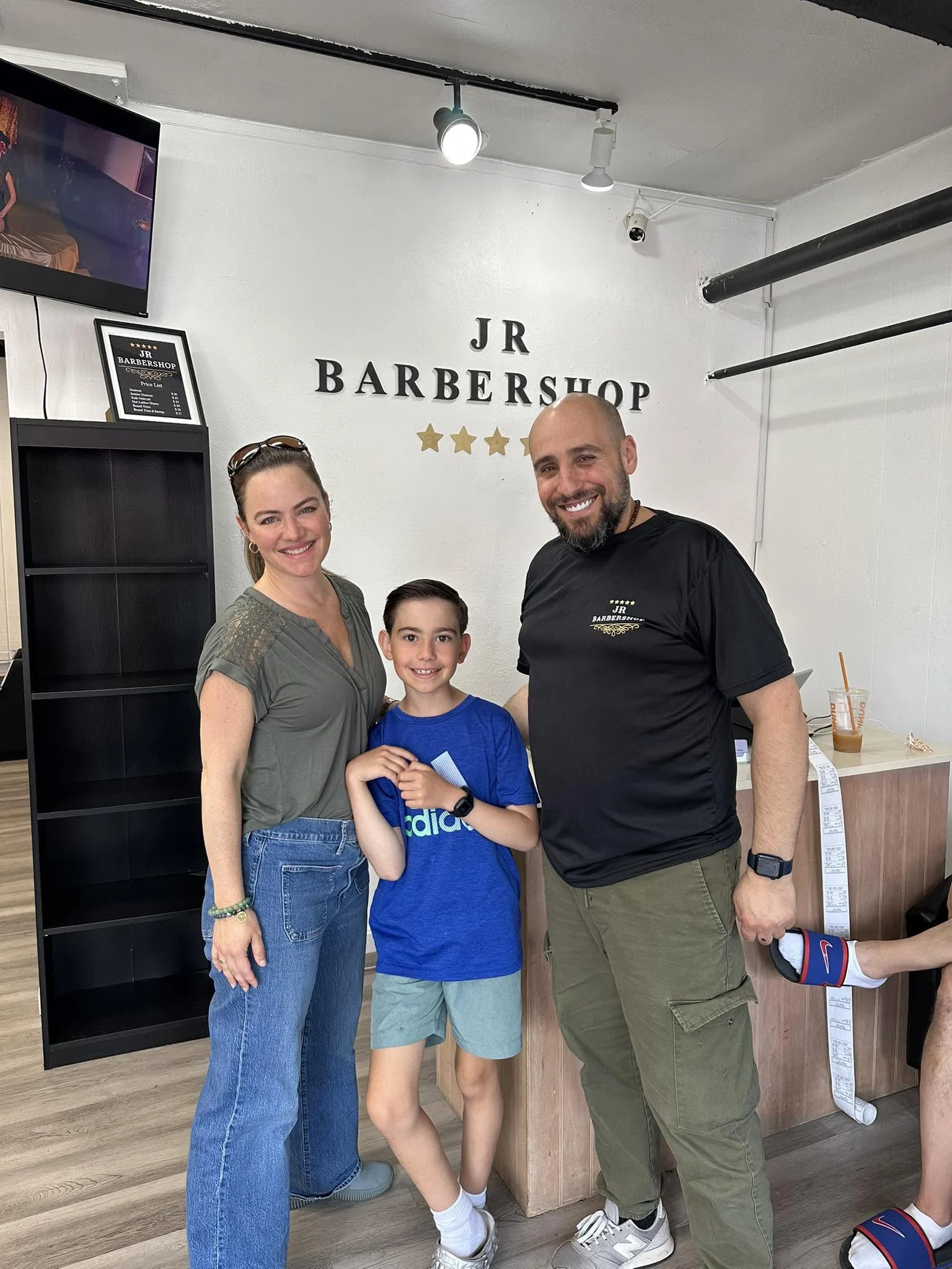 Barber and two visitors smiling together beneath the JR Barbershop wall sign